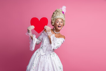 Graceful young woman dressed as a baroque aristocrat holding a red heart on a vibrant pink...