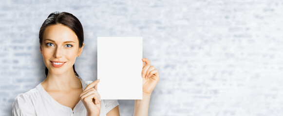 Smiling brunette hair business woman businesswoman wear white shirt show point blank empty banner mockup signboard. Advertise concept. Free copy space for ad text. Bricks wall background. Wide image