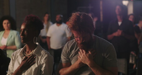 Man with hands on chest in deep prayer during spiritual gathering, surrounded by diverse congregation, African American woman praying beside him, faith and devotion emphasized