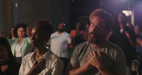 Obraz premium Man with hands on chest in deep prayer during spiritual gathering, surrounded by diverse congregation, African American woman praying beside him, faith and devotion emphasized