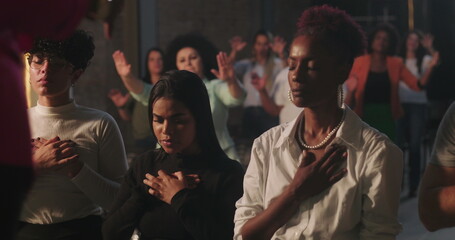 Congregation in prayer during spiritual gathering, hands on hearts, diverse group expressing faith and devotion, solemn worship moment with illuminated cross in background