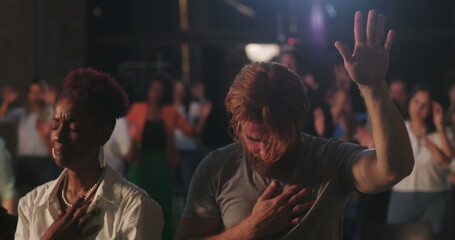 Congregation in prayer during spiritual gathering, hands on hearts, diverse group expressing faith and devotion, solemn worship moment with illuminated cross in background