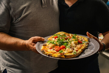 Freshly baked pizza topped with seafood, cherry tomatoes, and arugula, served on a plate held by two people, captured in a cozy indoor setting.