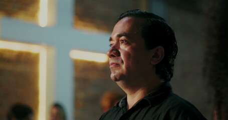 Middle-aged Latino man gazing forward with reflective expression in front of illuminated cross during spiritual gathering, showing deep connection and introspection