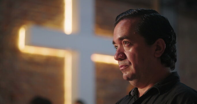 Close-up of man of Hispanic descent in church, illuminated cross in background, deep emotional reflection, faith and spirituality, peaceful moment of solemn introspection