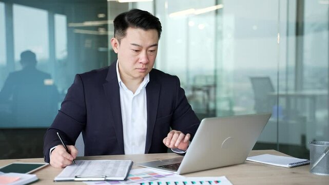Busy asian businessman fills out documents with a pen using laptop computer sitting at a work desk at workplace in business office. Financier doing paperwork, financial report, engaged in accounting