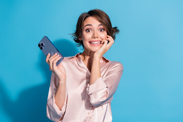 Young woman in pink satin blouse smiling joyfully, holding a smartphone against a vibrant blue...