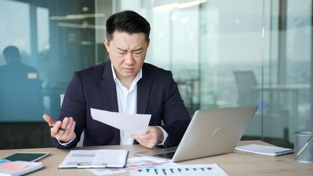 Frustrated asian businessman unhappy with financial results sitting at desk in business office. Upset puzzled financier reviewing documents and looking at laptop screen, unhappy with bad indicators