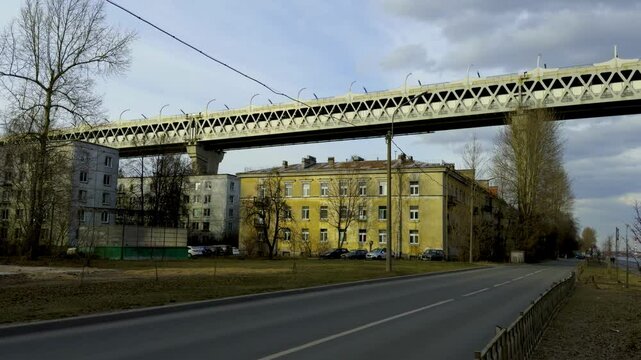 A wide-angle shot captures the industrial beauty of Kanonersky Island, showcasing the massive steel truss bridge spanning overhead. In the foreground, a paved road stretches alongside a row of