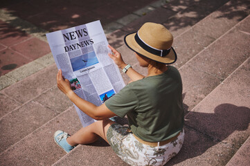Woman Reading a Newspaper Outdoors on Sunny Steps, Wearing Stylish Hat and Casual Clothes