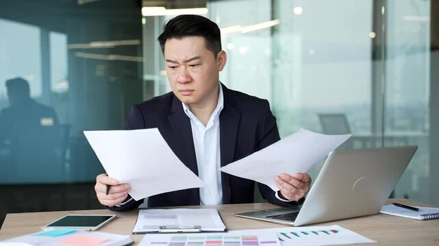 Confused disappointed asian businessman having difficulty with paperwork sitting at desk at workplace in business office. Puzzled worker in a suit looks at documents and cannot understand problem