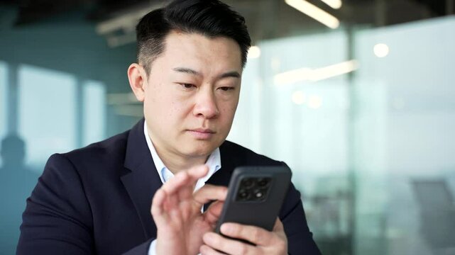 Serious asian businessman is using browsing mobile phone at workplace in business office. Entrepreneur in formal suit is chatting online with client or reading writing message on smartphone. Close up