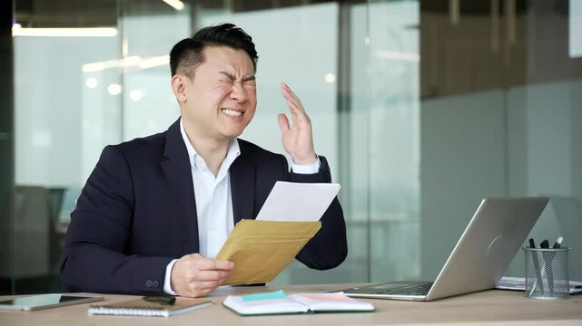 Worried asian businessman reading letter with bad news sitting at desk at workplace in business office. Disappointed upset entrepreneur in formal suit frustrated by received negative notification