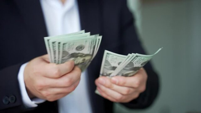 Close up. Male hands count one hundred dollar US bills. Asian businessman in formal suit counting banknotes while standing in modern business office. Man holds cash in hands. Financial success concept