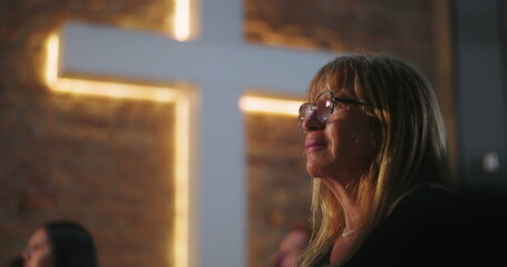 Woman with glasses looking up, emotional moment in front of illuminated cross at church during...