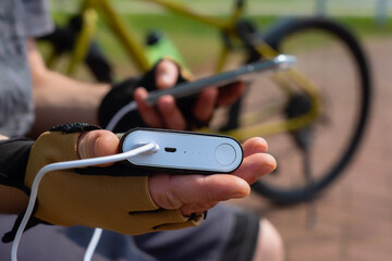 A male cyclist wearing sports gloves charges his smartphone with a portable power bank. The concept of modern technology and active lifestyle in nature, gadgets and energy on the go.