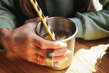 Woman holding metal mug. Striped straw in iced drink. Hand with ring and manicured nails. Cozy sweater in sunlight. Refreshing beverage moment. Casual drink on wooden table.