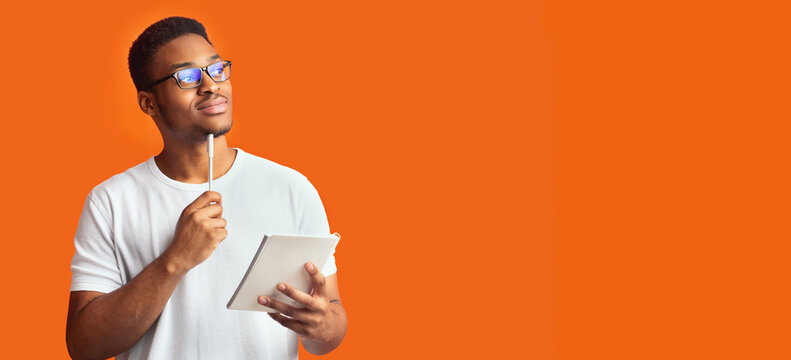 Pensive Redactor. Dreaming afro man in glasses taking notes, writing ideas, looking at copyspace, orange wall, panorama