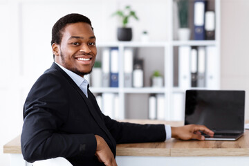 Successful african american businessman posing in his office, using modern laptop with blank...