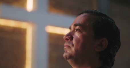 Close-up of man with emotional expression during worship in church, illuminated cross, reflecting...