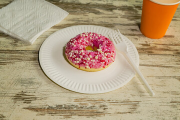 A donut with pink glaze on a disposable plate with a fork, next to a napkin and a glass in the background, close-up, background for a culinary blog mockup, donut on the table.