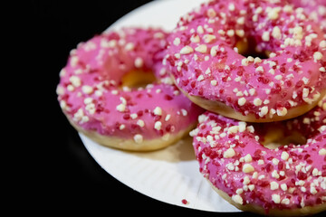 Doughnuts with pink glaze and culinary crumbs on a white plate on a black background, close-up, selective focus.