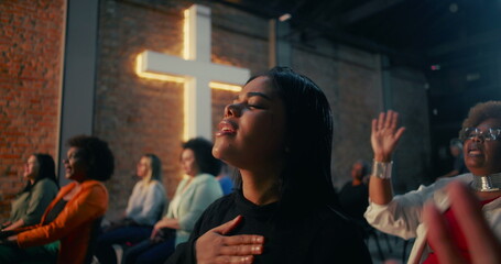 Hispanic woman of African descent with emotional expression, hands raised in worship as part of diverse congregation in Christian faith, cross in background