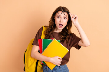 Happy young student holding colorful notebooks while adjusting glasses on beige background