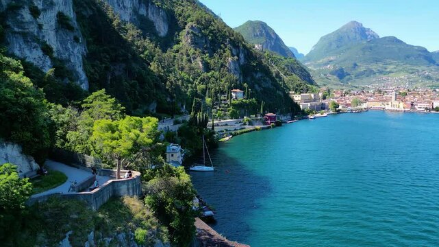 Aerial view of Riva del Garda with Torre Apponale, MAG museum, bike paths, boats and river ferries connecting towns, surrounded by mountains and clear Lake Garda water, full of tourists
