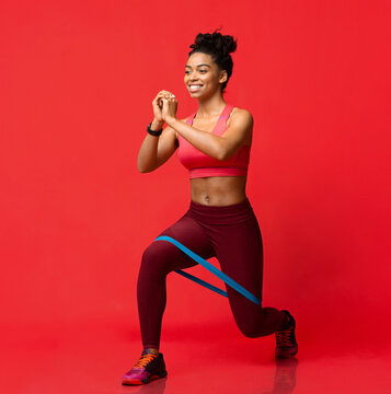 Cheerful black fitness girl making working out with resistance band over red studio background