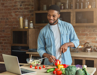 Happy african-american man preparing vegetable salad while looking on laptop