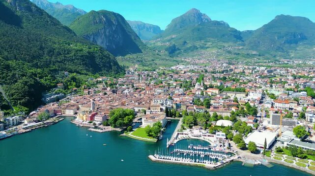 Aerial view of Riva del Garda with Torre Apponale, MAG museum, bike paths, boats and river ferries connecting towns, surrounded by mountains and clear Lake Garda water, full of tourists