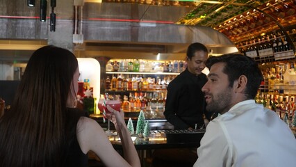 Bartender serving mixed punch cocktail Christmas theme garnished with candy cane serving to woman tasting drink on bar decorated green neon snow Christmas tree special festive couple time. Vinosity.