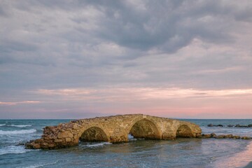 At the edge of the azure sea, an ancient stone bridge emerges from the waves as dusk casts warm hues across the horizon. Zakynthos summer tourism reveals captivating coastal charm
