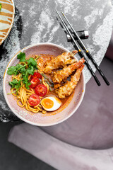A bowl of ramen with crispy tempura shrimp, halved boiled egg, cherry tomatoes, and fresh cilantro, served on a marble table with chopsticks, capturing the essence of Japanese cuisine