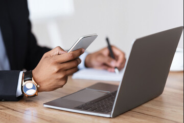 Unrecognizable black manager in suit holding smartphone, taking notes, sitting in front of laptop, office interior, cropped. African american businessman using modern gadgets for business