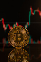 Vertical picture of a bitcoin standing on a reflective surface, with a stock chart in the background. Selective focus on the coin with slight blurred background.