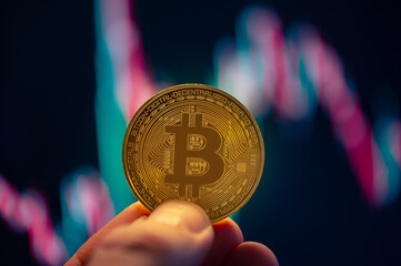 A person holding a bitcoin in front of stock chart with red an green candles. Selective focus on the coin, with a slight blurred background