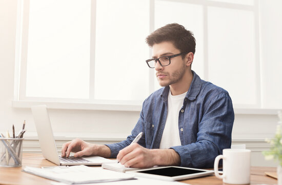 Fototapeta Young concentrated man typing on laptop and taking notes, in modern office. Handsome programmer working on computer and writing message, copy space