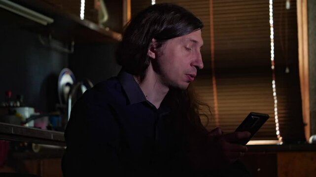 A mid-shot captures a curious mood as a man with long hair looks at his smartphone, with a distinct blue light reflecting on his face, suggesting a video call or content viewing in a dimly lit room
