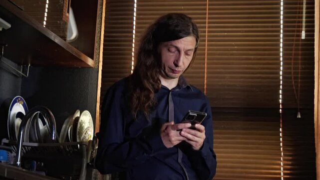 A mid-shot captures a curious mood as a man with long hair looks at his smartphone, with a distinct blue light reflecting on his face, suggesting a video call or content viewing in a dimly lit room