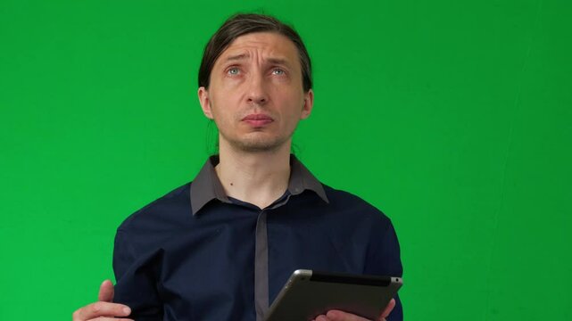 A mid-shot captures the tired and frustrated mood of a man holding a digital tablet, resting his hand on his cheek, against a vibrant green screen, ideal for customizable backdrops