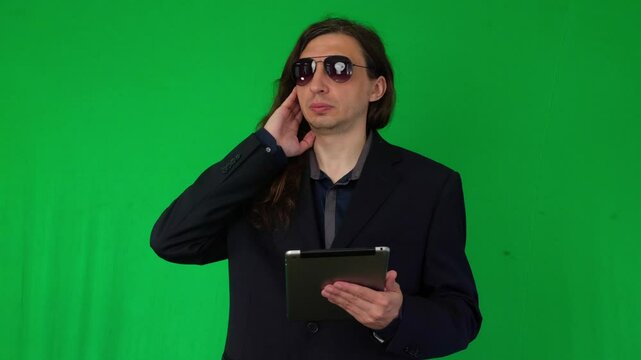 A mid-shot captures a serious and clandestine mood as a man in a suit and sunglasses, holding a tablet, touches his ear as if listening to an earpiece, against a green screen background