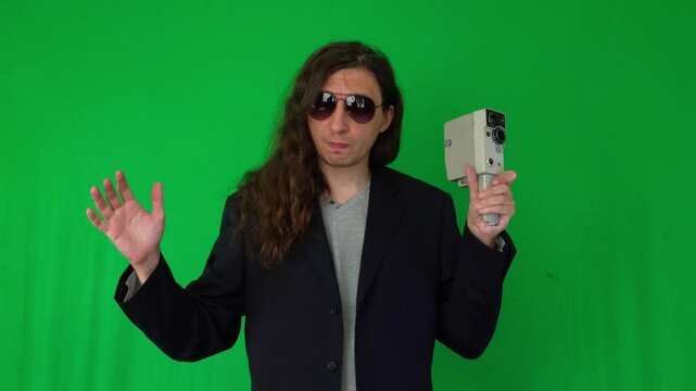 A mid-shot captures a retro and authoritative mood as a man in sunglasses holds a vintage film camera and makes a "stop" gesture, set against a vibrant green screen
