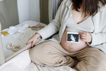 Pregnant woman holding ultrasound picture at baby bump and sitting at baby crib with mobile,  clothes and toy. Pregnancy. Baby room preparation