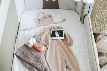 Pregnancy and getting ready for baby. Modern baby crib with stylish baby clothes, toy, boho mobile, ultrasound picture.Preparation and making nursery
