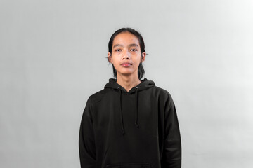 Portrait of a young Asian person standing against a plain gray background, wearing a black hoodie, looking directly at the camera with a neutral expression.