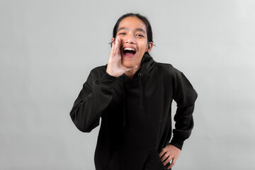 Asian man wearing a black hoodie shouting with hand near mouth, smiling and looking excited,...