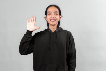 Asian man with long hair wearing a black hoodie smiling and raising his hand in a friendly greeting...