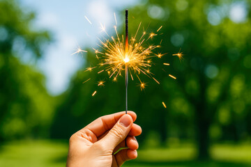 Fototapeta premium Close-up of a hand holding a lit sparkler in broad daylight with a lush green forest background, capturing a vibrant summertime celebration vibe with natural light and patriotic festivity